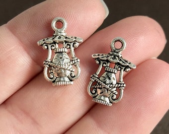 May include: Two silver-toned metal charms in the shape of a lamp with a decorative design. The charms have a loop at the top for attaching to a necklace or bracelet.