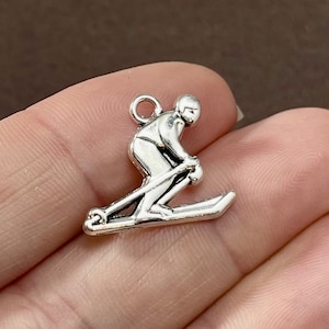 May include: Silver charm of a skier in a downhill skiing pose. The charm is detailed and has a loop for attaching to a necklace or bracelet.