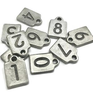 May include: A set of ten silver metal tags with black numbers 1 through 9 and 0. Each tag has a hole for hanging.