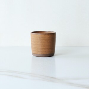 May include: A cylindrical ceramic cup with a warm, earthy brown tone and a matte finish. The exterior features subtle, horizontal ridged textures along the body, while the base is a darker, solid brown color with a smooth, unglazed appearance.
