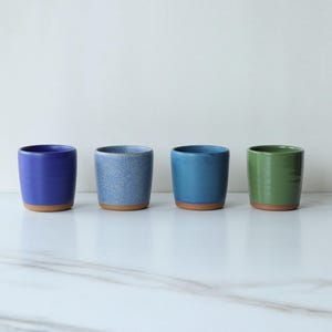 May include: Four ceramic tumblers in shades of blue and green, each with a natural clay-colored base. The tumblers are arranged in a row against a white background. The colors include a deep blue, speckled blue, teal, and a vibrant green.