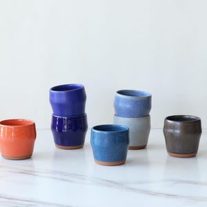 May include: A collection of six ceramic cups in various colors including orange, dark blue, light blue, grey, and brown. The cups feature a tapered shape with a smooth finish and a visible terracotta-colored base on each individual piece.