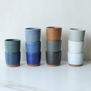 May include: A collection of ceramic cups in various colors, including blue, green, brown, gray, and white. Each cup is stacked, showcasing a minimalist design with a contrasting brown base. The cups are arranged on a white surface.
