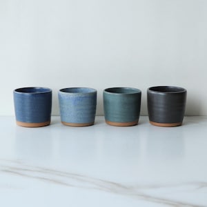 Puede incluir: Cuatro tazas de cerámica en tonos azules y grises dispuestas en fila. Cada taza tiene un acabado texturizado y una base natural sin esmaltar. Las tazas varían en color, desde un azul marino intenso hasta un gris antracita.