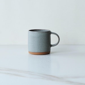 May include: A ceramic mug with a gray body and a brown base. The mug has a simple, cylindrical shape with a handle on the side. The mug is sitting on a white surface.
