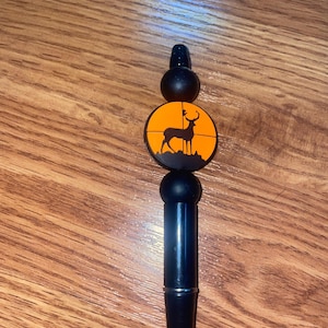 May include: Black pen with a deer silhouette design. The pen features a black body, a round black top, and an orange circle with a deer silhouette. The pen is designed for writing.