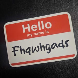 Puede incluir: Una etiqueta rectangular con esquinas redondeadas. La sección superior es roja con la palabra "Hello" en blanco, y "my name is" debajo. El nombre "Fhqwhgads" está impreso en negro en la sección blanca.