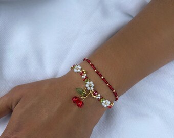 Op de afbeelding: Twee armbanden met kralen. Eén is een delicate rode kralenarmband. De andere is een goudkleurige armband met witte en rode kralen en een kersenbedel met rode kersen en groene bladeren.