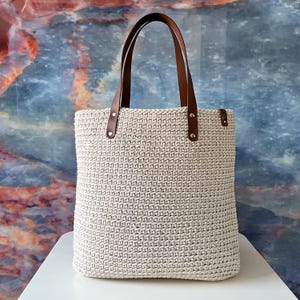 Könnte beinhalten: Eine cremefarbene, gehäkelte Tragetasche mit braunen Ledergriffen und Akzenten. Die Tasche ist rechteckig mit einer strukturierten Oberfläche und einem schlichten, eleganten Design. Die Griffe sind mit Nieten befestigt.