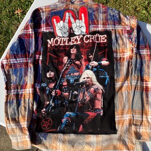 以下が含まれることがあります： 青とオレンジの格子柄のフランネルシャツで、背面に大きな「Motley Crue」のパッチが付いています。パッチにはバンドの写真とバンド名が白い文字で書かれています。
