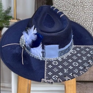 May include: A navy blue fedora hat with a wide brim. The hat features a patchwork design with a patterned fabric section and is embellished with light blue feathers and a silver-toned chain. The hat is sitting on a wooden chair.