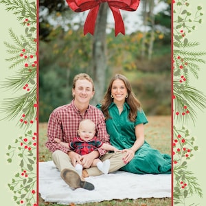 May include: A Christmas card featuring a family of three, a man, a woman, and a baby, sitting on a white blanket in a grassy field. The family are smiling and looking at the camera. The card has a green background with a red bow at the top and the text "Merry Christmas" at the bottom.