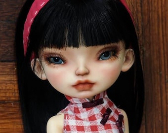 May include: A doll with black hair, blue eyes, and a red and white checkered dress. The doll has a red bow headband and a matching bow on the dress. The doll's arms and legs are articulated.