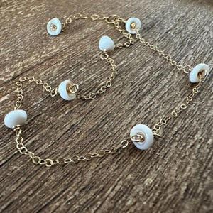 Peut inclure: Un collier délicat en chaîne dorée avec de petites perles blanches et rondes espacées régulièrement le long de sa longueur. Le collier est présenté sur une surface en bois, mettant en valeur son design élégant et son savoir-faire.