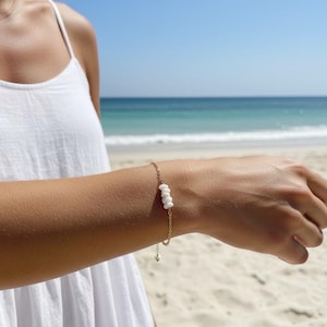 Peut inclure: Un bracelet à chaîne dorée orné de quatre perles blanches ovales. Le bracelet est porté sur un poignet, avec une plage et un océan en arrière-plan. La personne porte une robe blanche.