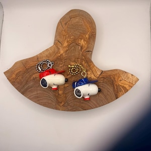 May include: Two Snoopy-themed keychains, one with a red baseball cap and the other with a blue cap, rest on a decorative wooden slab. The keychains are white with black ears and eyes, and red accents. The wooden slab has a unique, natural grain.