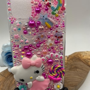 May include: A clear phone case decorated with pink beads, stars, and a rainbow. Features a Hello Kitty figure, a lollipop, and the name "ALEXA". The case is embellished with various pink and white accents.