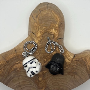 May include: Two character keychains: a white Stormtrooper helmet and a black Darth Vader helmet. Both keychains are attached to silver ball chains. The keychains are displayed on a wooden, organic-shaped surface.