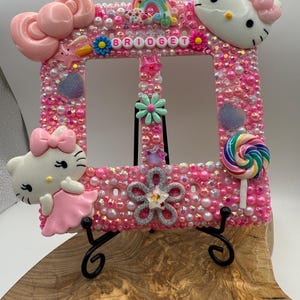 May include: A pink, bedazzled picture frame with two openings, decorated with Hello Kitty figures, bows, and a rainbow. The name "BRIDGET" is spelled out across the top. The frame is supported by a black metal stand.