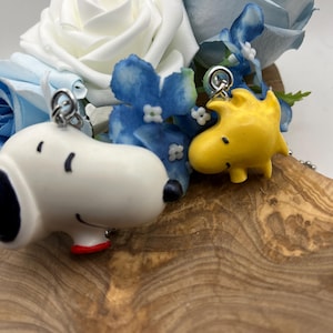 May include: A Snoopy and Woodstock keychain set. The Snoopy keychain is white with black and red accents. The Woodstock keychain is yellow. The keychains are displayed with blue and white flowers on a wooden surface.