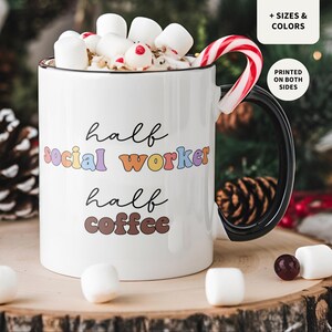 Peut inclure: Une tasse en céramique blanche avec une anse noire. La tasse porte l'inscription "half social worker half coffee" en lettres colorées. La tasse est remplie de chocolat chaud et de guimauves. Une canne en sucre est posée contre la tasse.