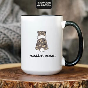 May include: White ceramic mug with a black handle and rim. It features a cartoon illustration of an Australian Shepherd dog from the rear, with the words "aussie mom" in a script font below. The mug is set on a wooden surface.