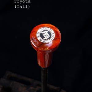 Könnte beinhalten: Nahaufnahme eines hölzernen Schaltknaufs mit roter Oberfläche. Der Knauf hat ein silbernes Toyota-Logo und japanischen Text, der "Toyota - Synchro - Gear" lautet.