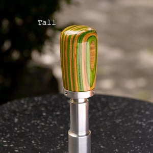 Może przedstawiać: Drewniana gałka zmiany biegów z paskiem w kolorze zielonym, brązowym i beżowym. Gałka jest przymocowana do srebrnej metalowej podstawy. Słowo "Tall" jest wydrukowane na zdjęciu na biało.