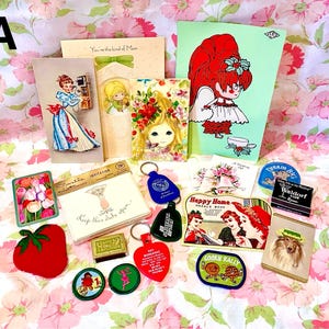 May include: A collection of vintage cards, keychains, and other ephemera, including a card with the text "You're the kind of Mom", a card with a red-haired girl, a card with a floral design, a card with a strawberry, a card with a needle book, a card with a cookie rally, a card with a dog, and a card with the text "Dream Big".