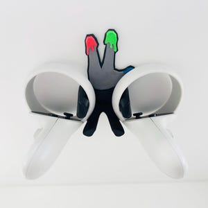 May include: A black wall-mounted controller holder shaped like a hand with two fingers extended. The fingertips feature red and green paint drips, and the holder supports two white virtual reality controllers with circular tracking rings.