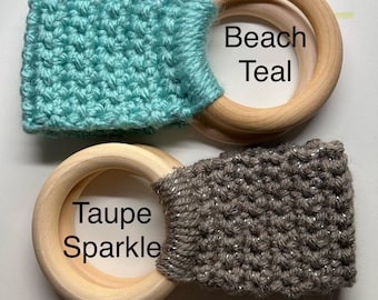 Puede incluir: Dos anillos de dentición de madera con tela de crochet unida. El anillo superior tiene una tela de crochet de color turquesa con el texto "Beach Teal". El anillo inferior tiene una tela de crochet de color topo con el texto "Taupe Sparkle".
