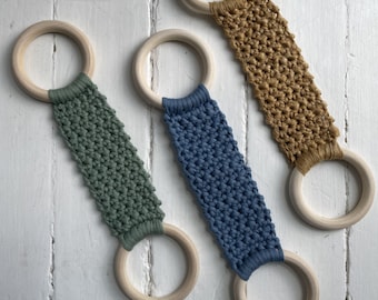 Puede incluir: Tres anillos de madera con correas de tejido de crochet en verde, azul y marrón. Los anillos cuelgan de una superficie de madera blanca.