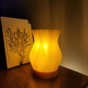 Puede incluir: Una lámpara de cerámica amarilla con un diseño ondulado. La lámpara está encendida y proyecta un brillo cálido.