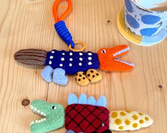 May include: Two handmade felt dinosaur keychains. One is blue with white dots and orange accents, the other is green and red with a yellow tail. Both have open mouths and small felt feet. An orange and blue keychain loop is attached to the blue dinosaur.