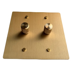 以下が含まれることがあります： 2つの丸い真鍮製のノブが付いた、ブラッシュゴールドの金属製ライトスイッチプレートです。