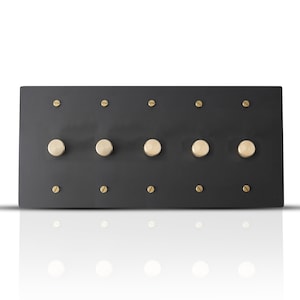 Op de afbeelding: Een rechthoekige, matzwarte wandlichtschakelaarplaat met vijf grote, ronde, goudkleurige dimmerschakelaars. Kleine gouden schroeven bevestigen de schakelaars en de plaat aan de muur. De plaat wordt weerspiegeld in het witte oppervlak eronder.