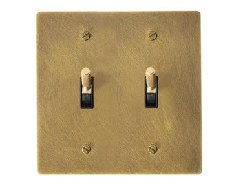 May include: A square, brass-colored double light switch plate with two black toggle switches. The plate is secured with four screws, one in each corner. The finish has a textured appearance.