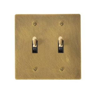 May include: A square, brass-colored double light switch plate with two black toggle switches. The plate is secured with four screws, one in each corner. The finish has a textured appearance.