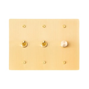 May include: A brass wall plate with three toggle switches and a round knob. The plate has a brushed finish and is mounted on a white wall.