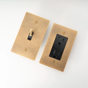 May include: Two brushed brass electrical wall plates, one with a single toggle switch and the other with a standard outlet. Both plates have a distressed finish and black accents.