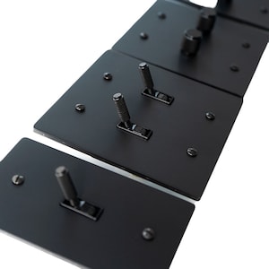 Puede incluir: Primer plano de varias placas de interruptores de luz de metal negro. Cada placa tiene un interruptor basculante texturizado y está asegurada con tornillos pequeños. Los interruptores están dispuestos en fila sobre un fondo blanco, destacando su diseño elegante y moderno.