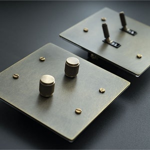 Può includere: Due placche per interruttori in ottone antico. Una placca ha due interruttori a levetta, l'altra ha due manopole rotanti. Ogni placca ha quattro viti in ottone. Le placche sono quadrate e hanno una finitura anticata.