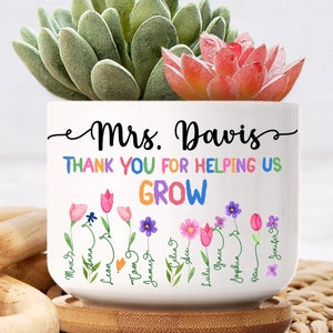 May include: White ceramic planter with the text "Mrs. Davis Thank You for Helping Us Grow" and a row of colorful flowers with names written on the stems. The planter is sitting on a wooden base.