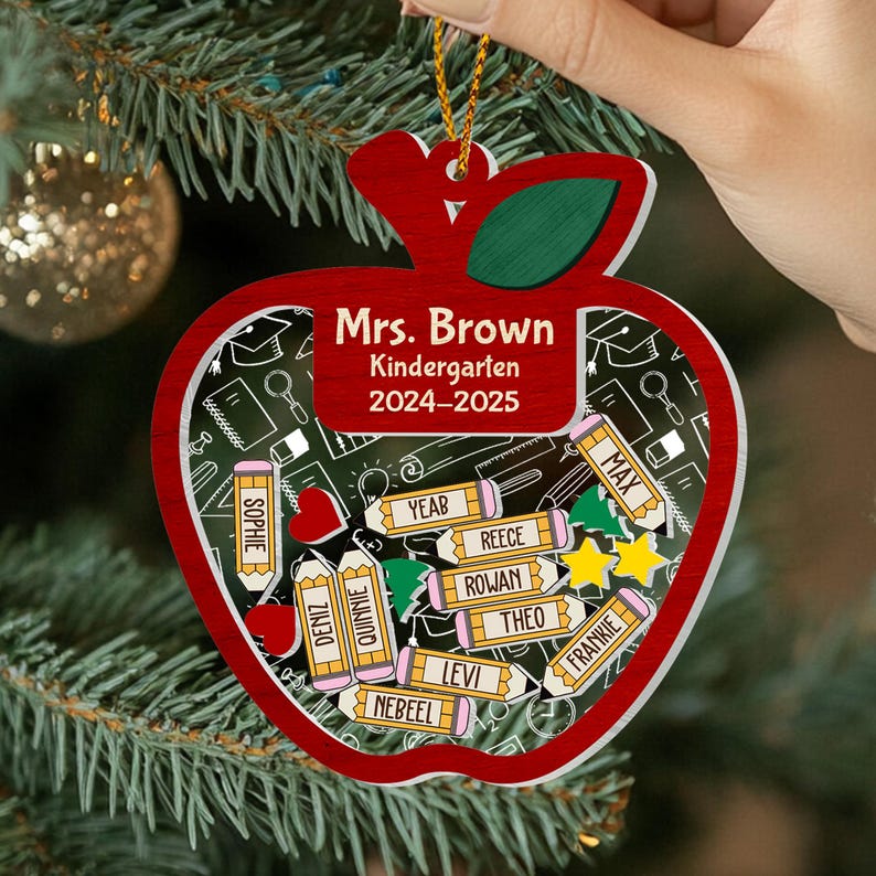 May include: A red apple-shaped ornament with a green leaf and gold string. The ornament has a clear center with a chalkboard design and pencils with names. Text reads "Mrs. Brown Kindergarten 2024-2025".