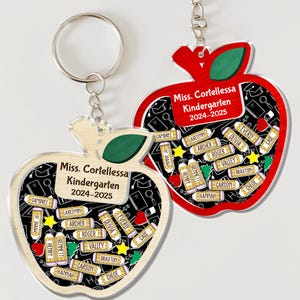 May include: Two apple-shaped keychains with a red and light wood grain design. Each keychain features the text "Miss. Cortellessa Kindergarten 2024-2025" and student names. The keychains have a silver keyring.