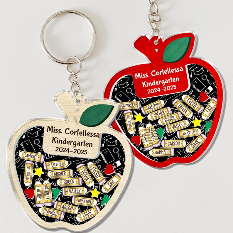 May include: Two apple-shaped keychains with a red and light wood grain design. Each keychain features the text "Miss. Cortellessa Kindergarten 2024-2025" and student names. The keychains have a silver keyring.