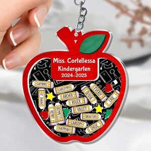 May include: A red apple-shaped keychain with a green leaf and a silver chain. The keychain features the text "Miss. Cortellessa Kindergarten 2024-2025" and student names on pencil-shaped tags. The background is black with white school-themed illustrations.