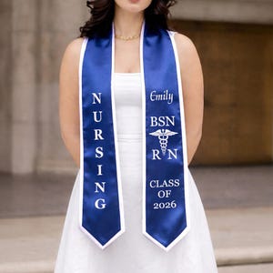 May include: A navy blue and white graduation sash with the word "NURSING" vertically printed on one side. The other side displays "Emily", "BSN", the RN symbol, and "CLASS OF 2026". The sash is worn over a white dress.