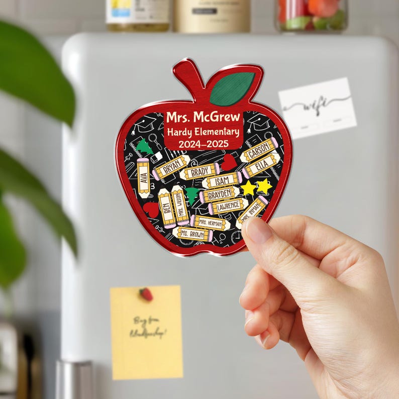 May include: A red apple-shaped magnet with a green leaf and a black interior. The magnet has the text "Mrs. McGrew Hardy Elementary 2024-2025" and student names. The magnet is being held up against a white refrigerator.