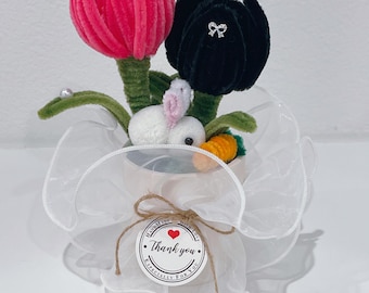 May include: A small white ceramic pot with a pink and black felt flower arrangement. A white bunny and a carrot are nestled in the pot. The pot is wrapped in white tulle and has a handmade with love tag that says "Thank you" with a red heart.